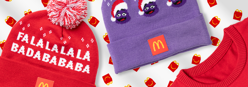 McDonald's-themed winter apparel on Happy Meal wrapping paper: red knit hat with 'FALALALALA BADABABABA' text, purple beanie with Grimace and logo patch, and red ribbed sweater.