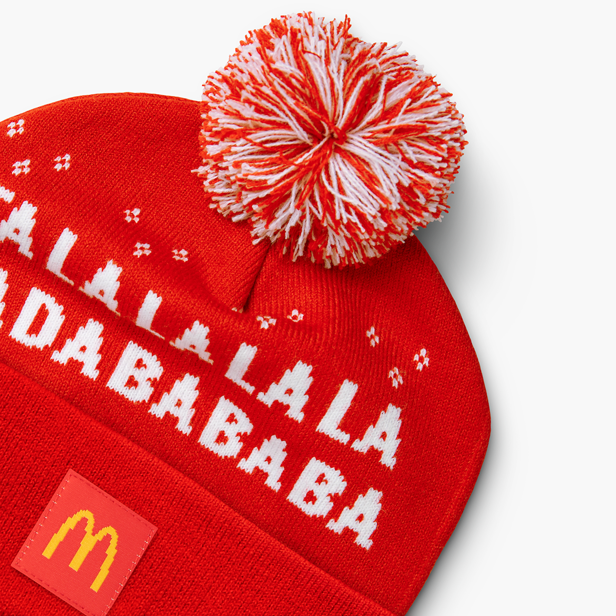 Front close-up of beanie with red-and-white pom, pixel-style text reading “Fa La La La La Ba Da Ba Ba Ba,” and McDonald’s patch on the cuff. 