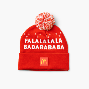 Front of red and white holiday pom beanie with white snowflakes, bold white text on the front, and a small McDonald’s Golden Arches patch on the cuff.