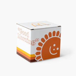 The mug next to a matching box featuring the same orange smiling sun